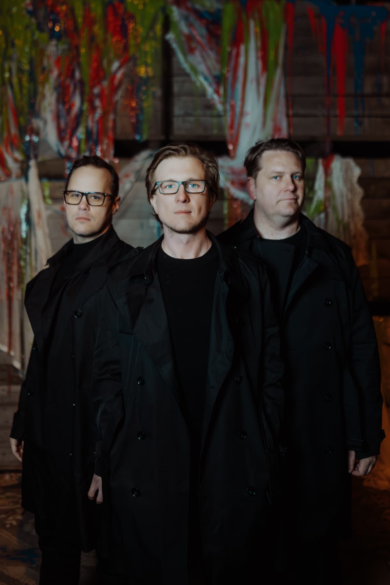 Trio Confusion Project w czarnych płaszczach na tle ściany pokrytej kolorową farbą, fot. Sisi Cecylia